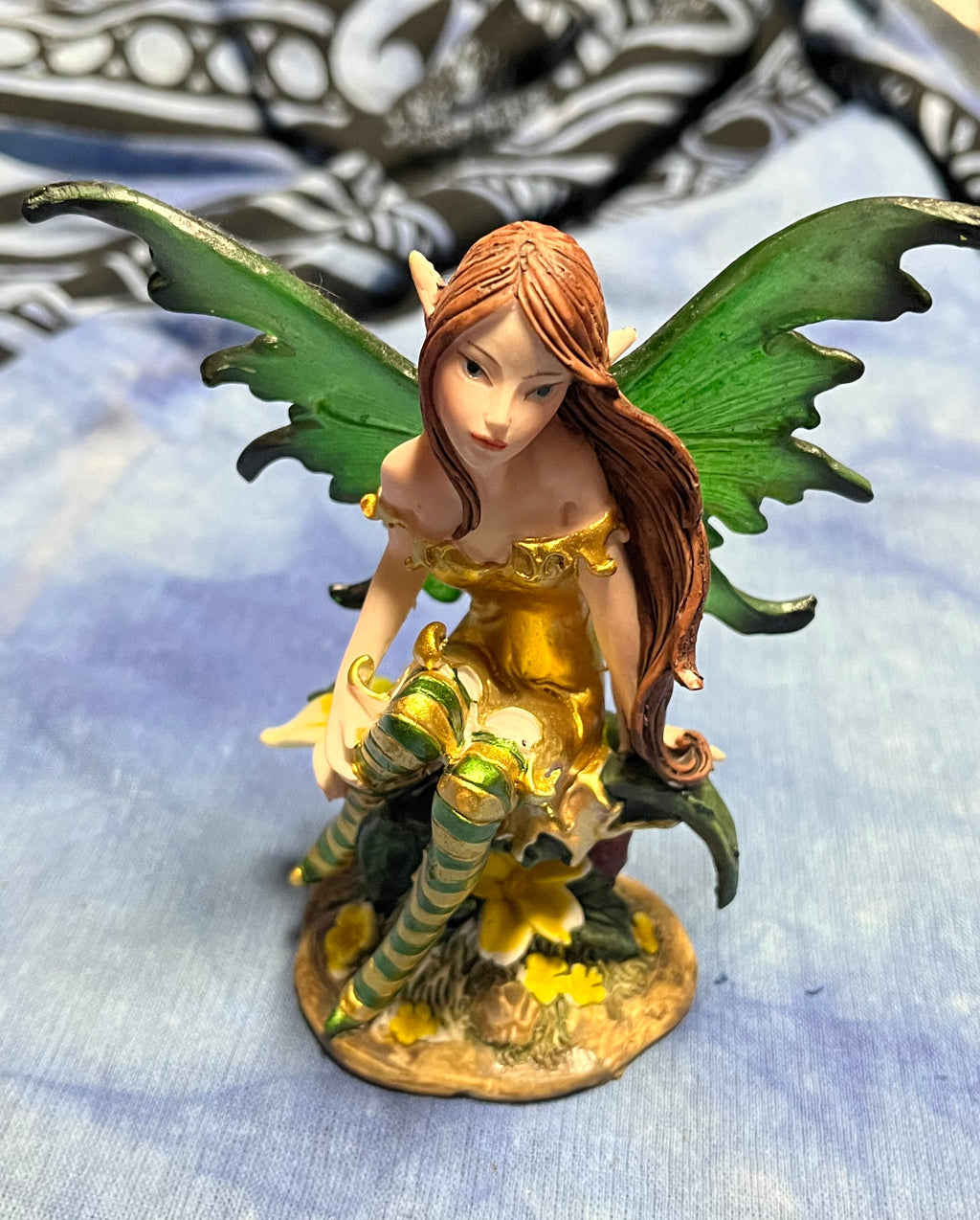 Fairy figurine with green wings and a gold dress holding flowers on a textured blue background